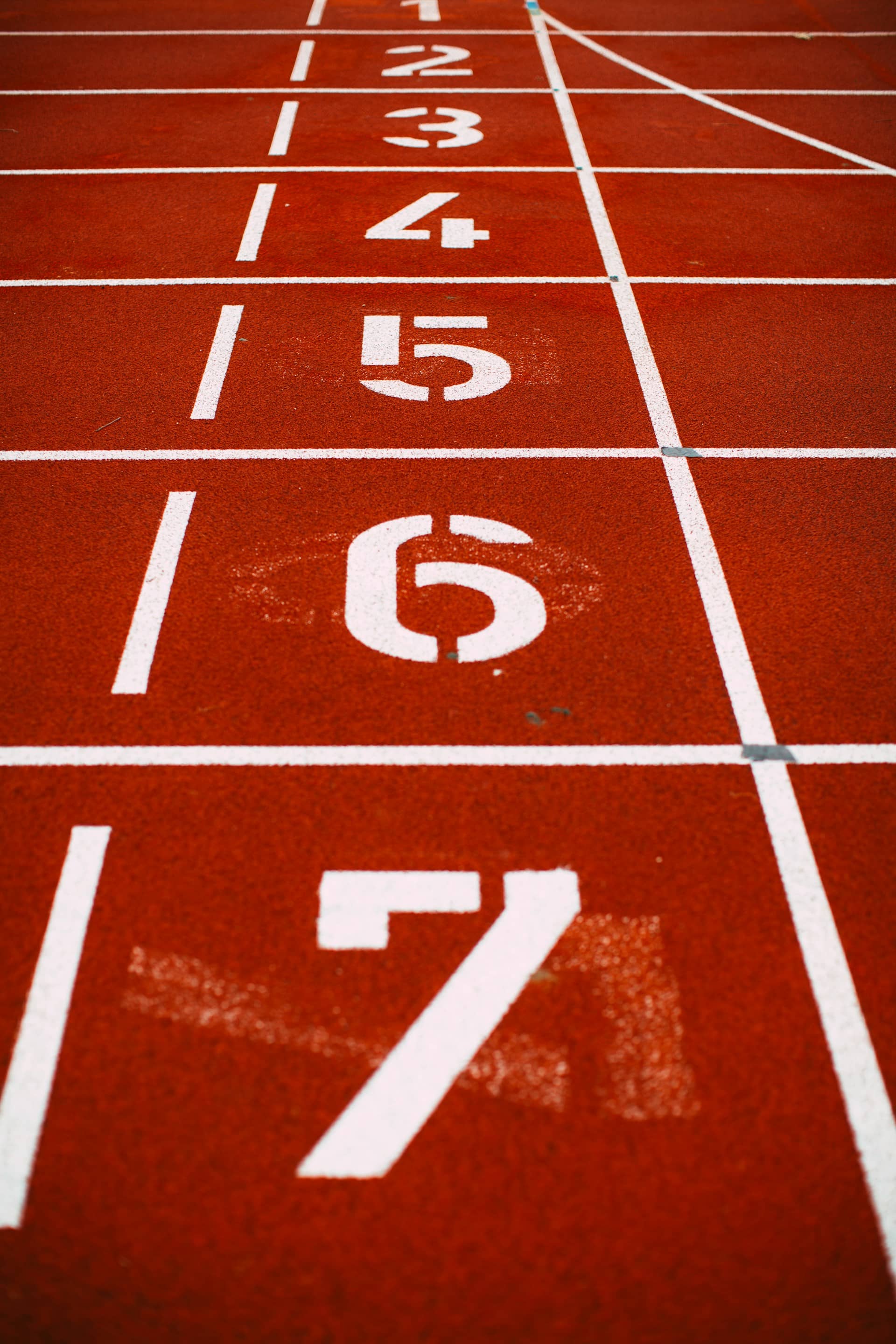 Numbered lane markings on a red running track, counting down from seven in the foreground toward the starting line